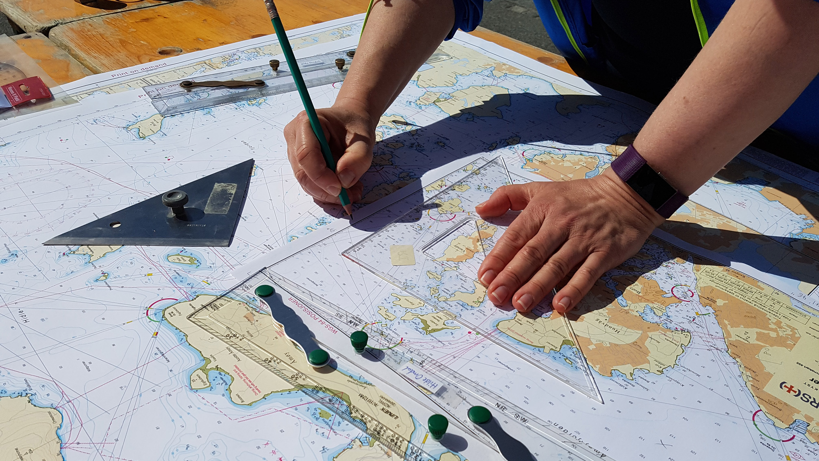 Et skolekart (sjøkart) ligger oppå et hovedseriekart. To hender plotter inn kurs med blyant og linjal. Foto.