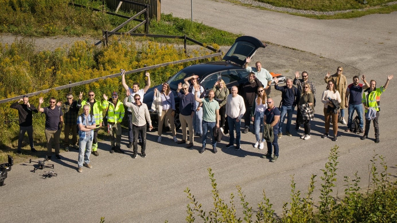 Deltakerne st&aring;r og vinker og smiler opp til en drone (ikke synlig) som tar bildet
